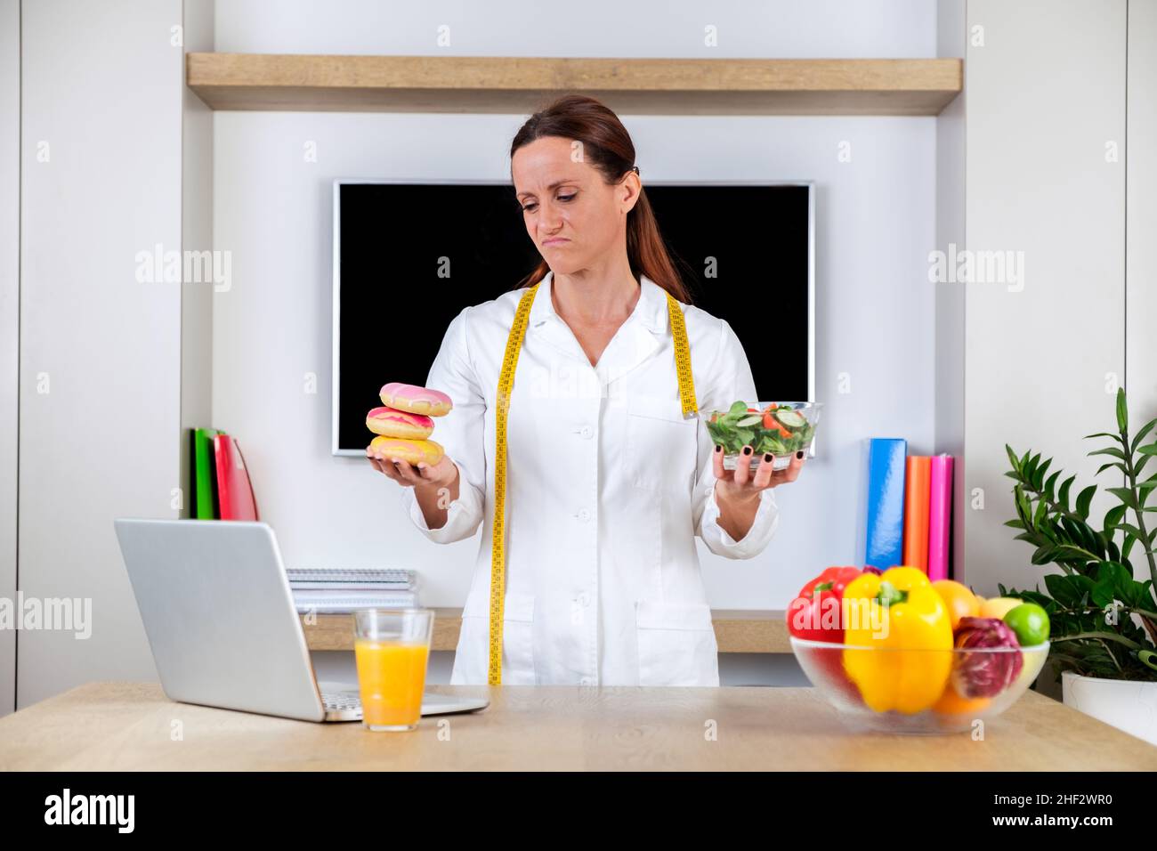 Jeune femme portant un uniforme nutritionniste tenant une salade et des beignets avec des émotions tristes dans une salle de bureau moderne et lumineuse.Choisir entre des aliments sains Banque D'Images
