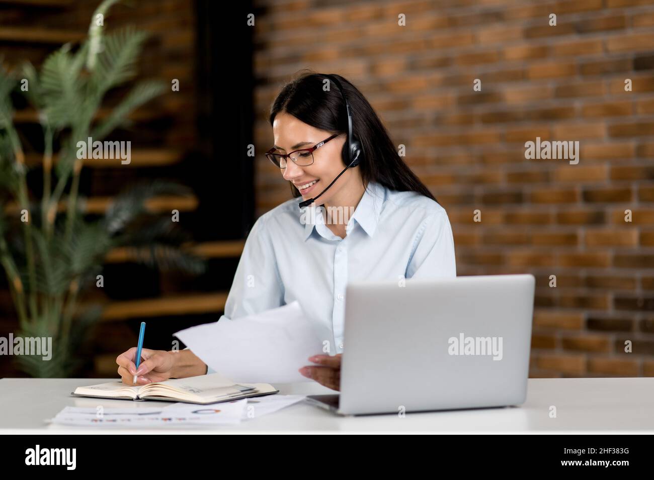 Femme caucasienne amicale, portant des lunettes et un casque, responsable, employé de soutien, consultant en ligne, opérateur,assis à l'ordinateur portable dans le bureau, enregistre l'information, mène une consultation en ligne Banque D'Images
