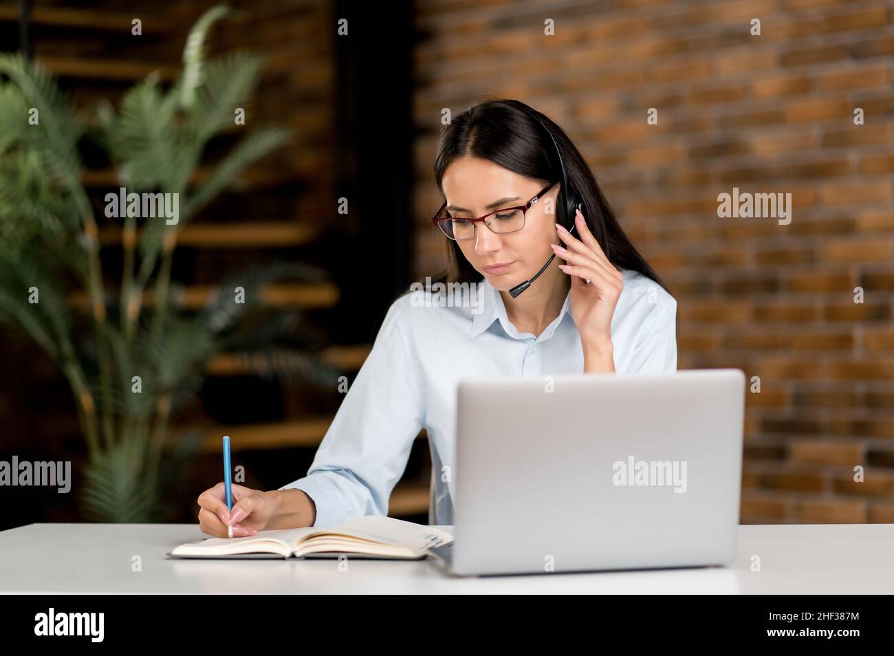 Femme caucasienne sérieuse, portant des lunettes et un casque, responsable, employé de support, consultant en ligne, opérateur,s'asseoir à l'ordinateur portable dans le bureau, enregistre l'information, mène une consultation en ligne Banque D'Images