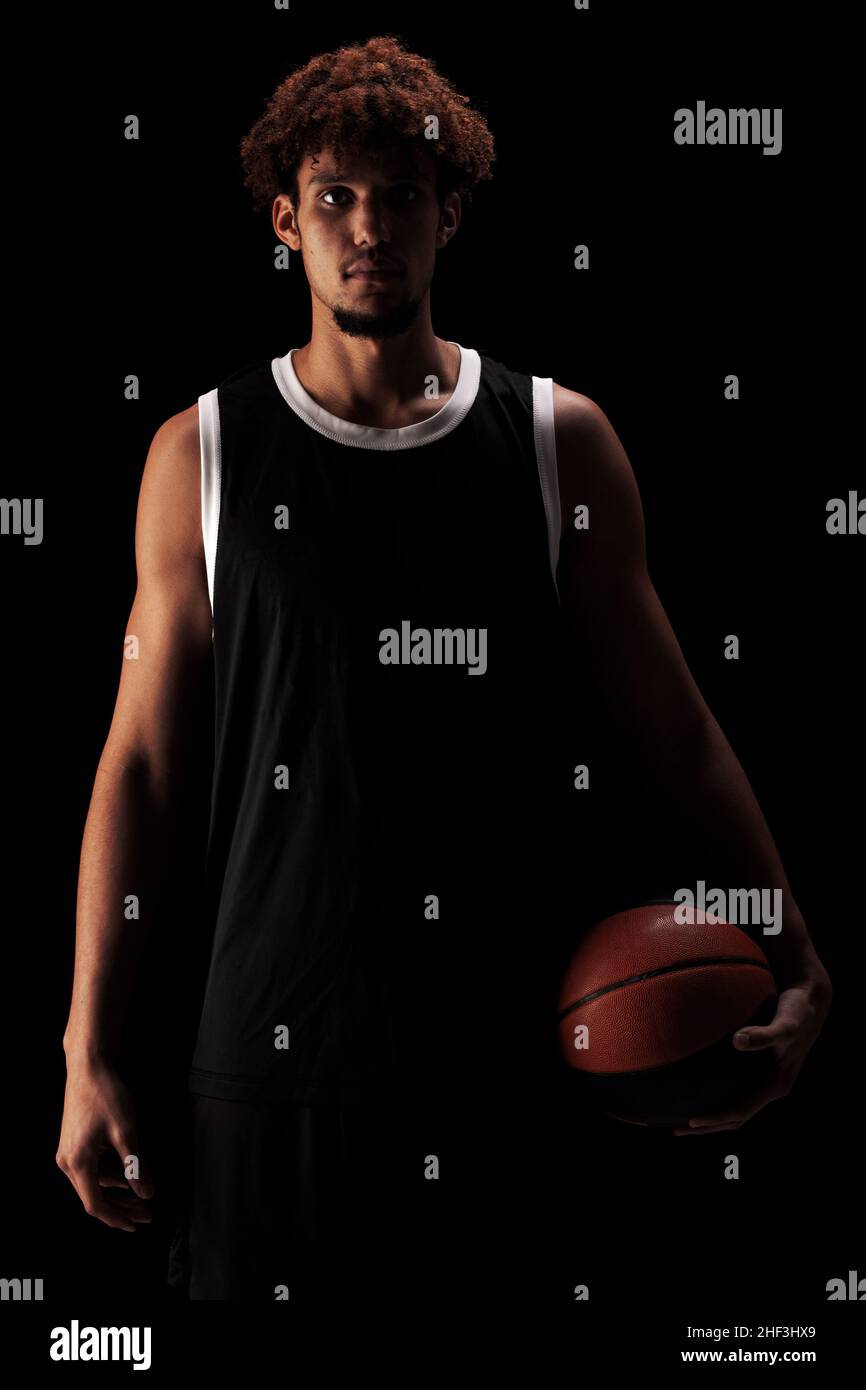 Joueur de basket-ball professionnel tenant une balle sur fond noir. Sérieux homme afro-américain concentré en uniforme sportif. Banque D'Images