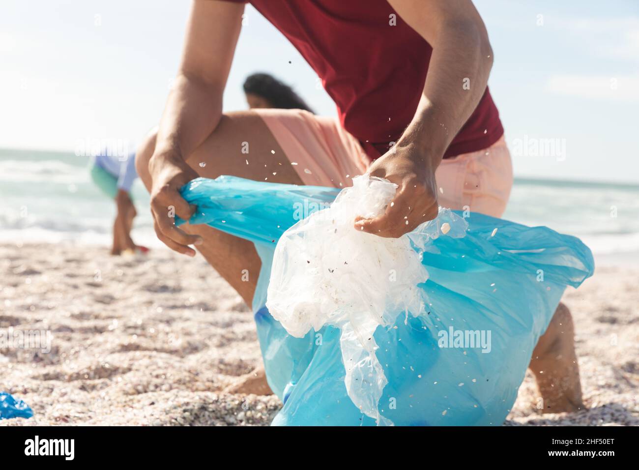 La partie basse de l'homme biracial collectant les déchets de plastique dans le sac à la plage le jour ensoleillé Banque D'Images