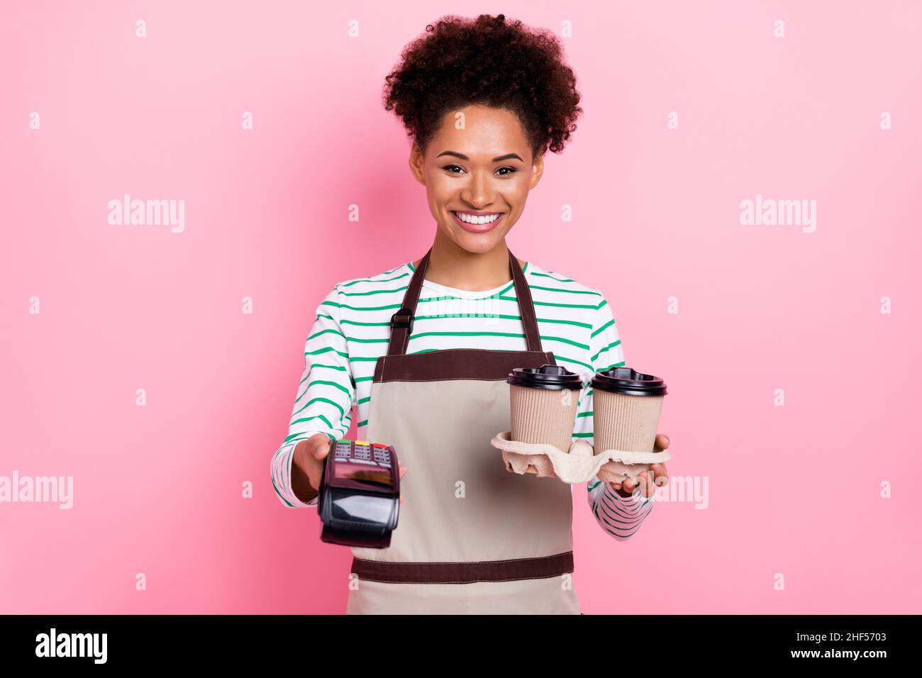 Photo de la jolie caissière gaie fille tenir café barista demande paiement porter tablier rayé chemise isolée couleur rose fond Banque D'Images