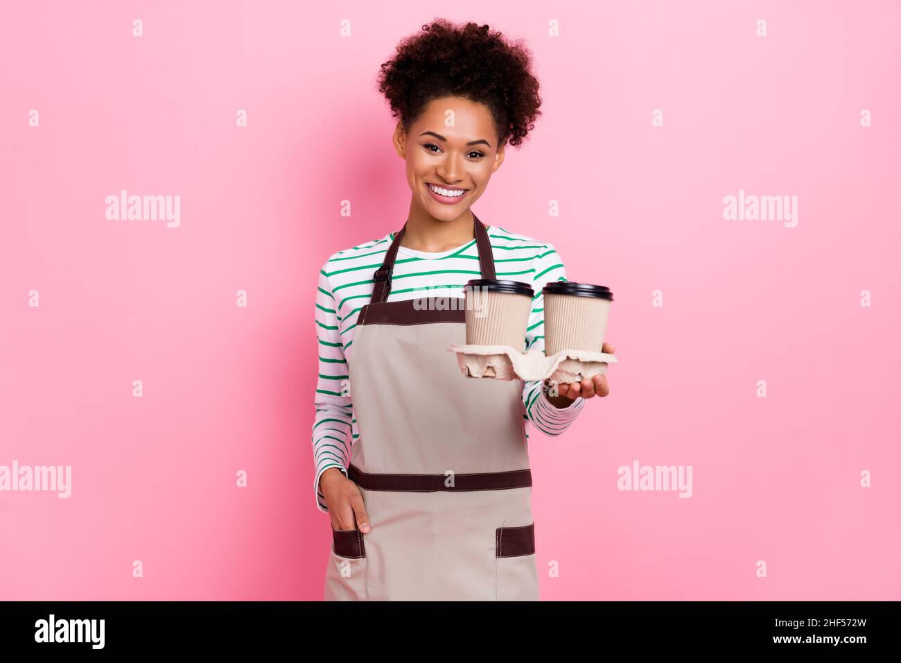 Photo de jolie fille positive tenir tasses de café de barista à emporter porter tablier rayé chemise isolée couleur rose fond Banque D'Images