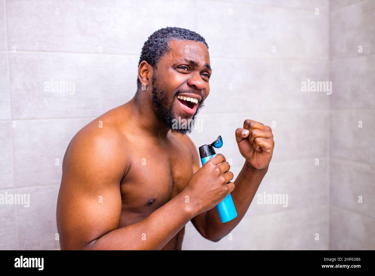 Beau jeune homme gai chantant en se lavant dans la douche, tenant une bouteille de shampooing comme un microphone Banque D'Images