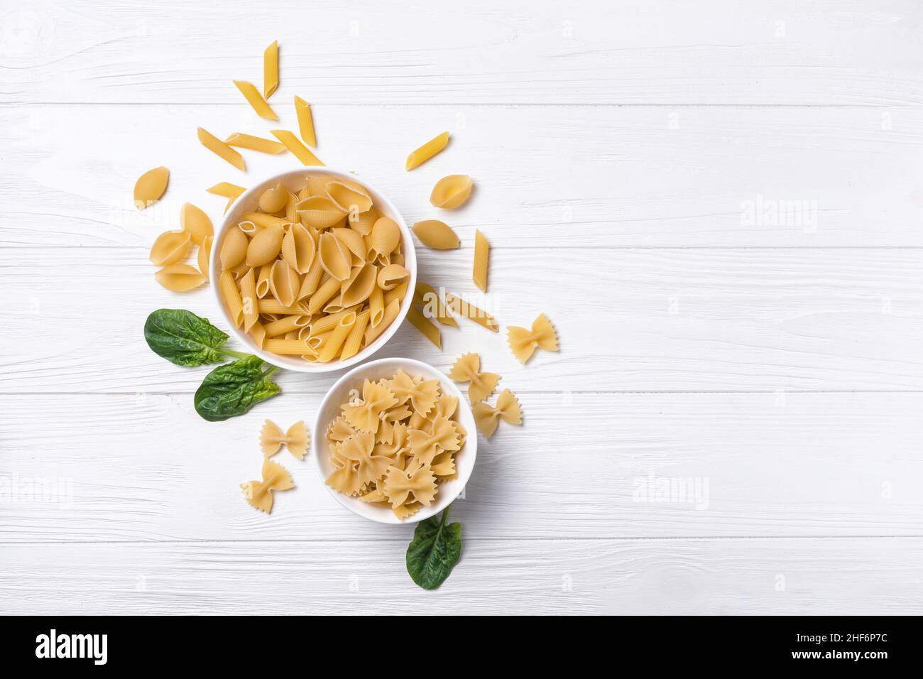 Vue de dessus sur une variété de pâtes italiennes crues dans des bols blancs avec des feuilles d'épinards verts frais sur fond de bois blanc Banque D'Images