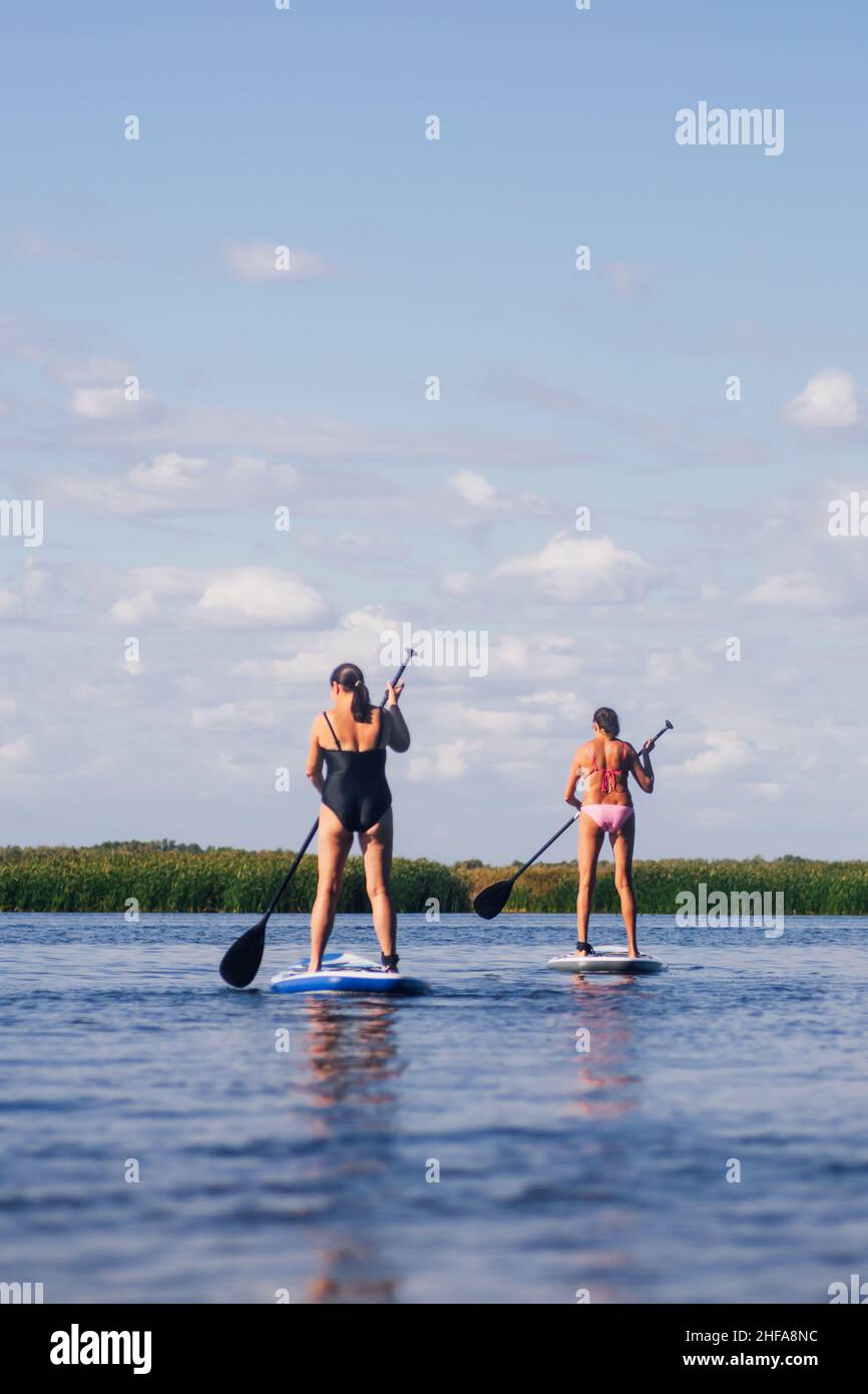 Faible angle des femmes plus âgées sur les planches de sup les deux oars en mouvement de la même manière sur le lac bleu avec des ondulations avec des roseaux verts en arrière-plan portant la natation Banque D'Images