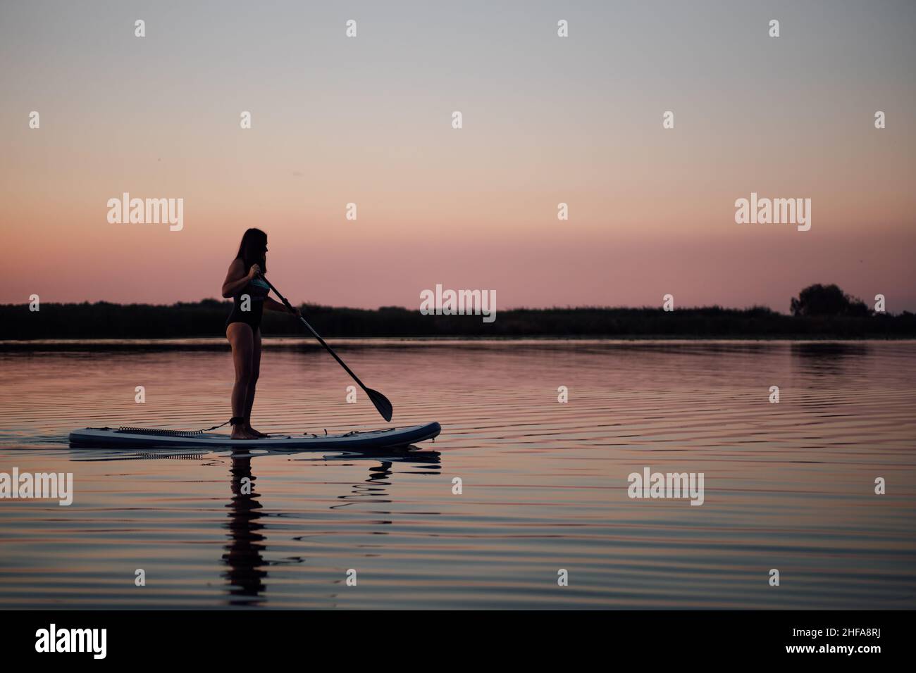 Femme d'âge moyen avec des mains rames droit sur le lac du soir avec ciel rose en arrière-plan en maillot de bain en été.Un style de vie actif pour Banque D'Images