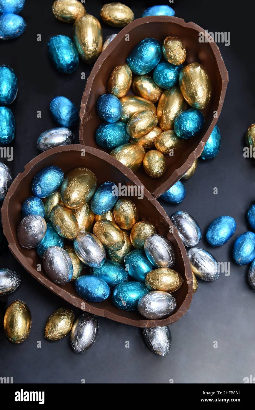 Pile de différentes tailles d'oeufs de pâques au chocolat enveloppés d'une feuille colorée en bleu, argent et or avec deux moitiés d'un gros oeuf de chocolat. Banque D'Images