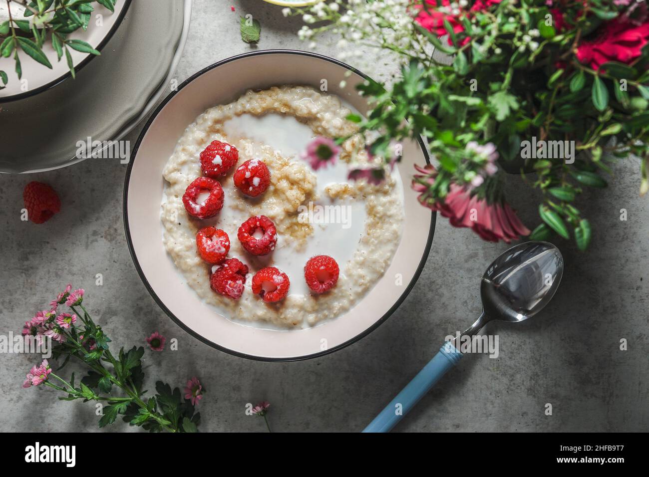 Bol de porridge maison avec framboises sur table de cuisine avec cuillère et fleurs.Idée de petit déjeuner végétalien sain à la maison.Vue de dessus. Banque D'Images