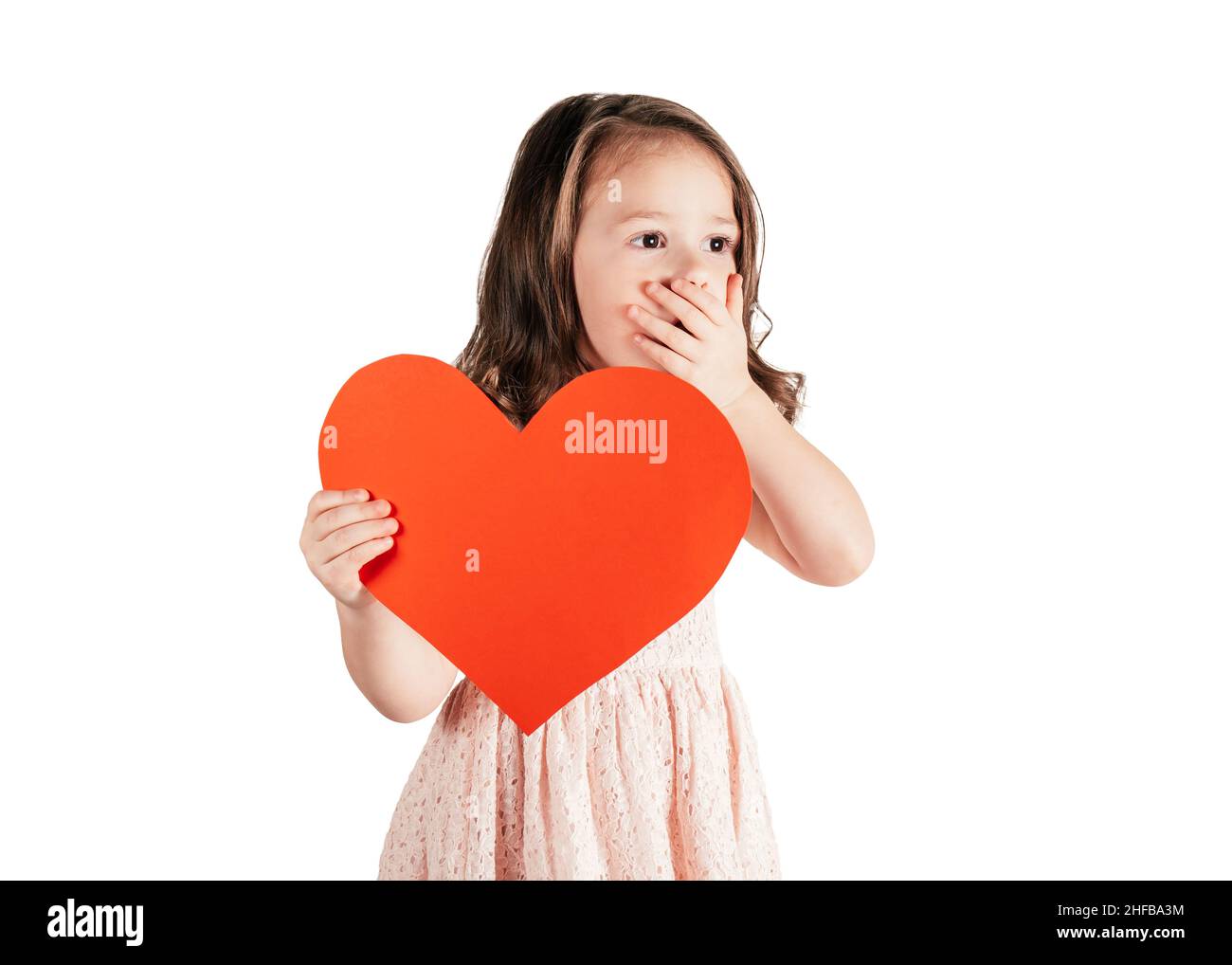 Portrait de petite fille avec les cheveux longs bouclés tenir dans la main rouge papier coeur mettant la main sur la bouche isolée sur fond blanc.Félicitations, Valen Banque D'Images