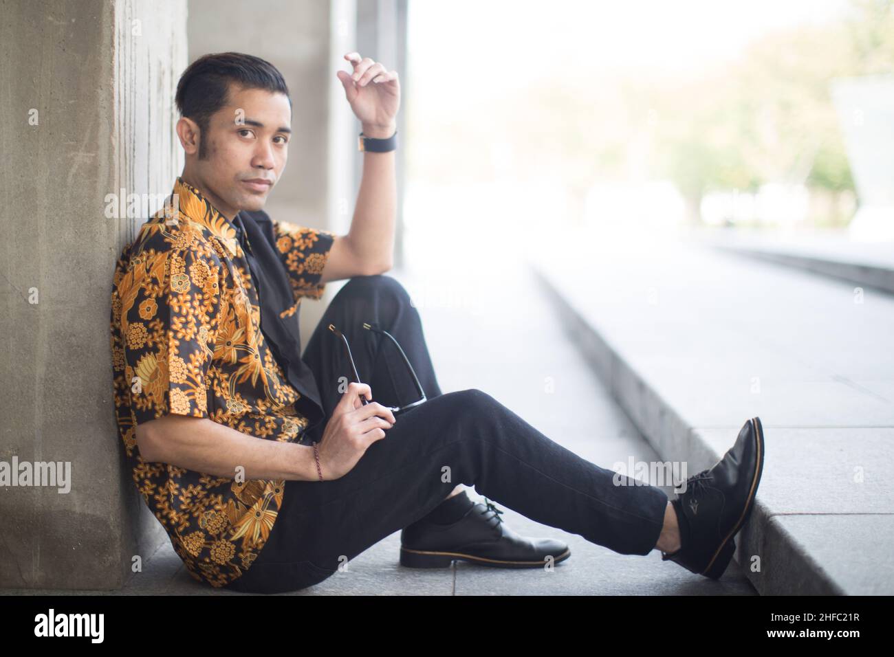 Un modèle masculin vêtu de Batik indonésien est assis et tient des lunettes de soleil sur fond de piliers au long Museum, West Bund, Shanghai, Chine Banque D'Images