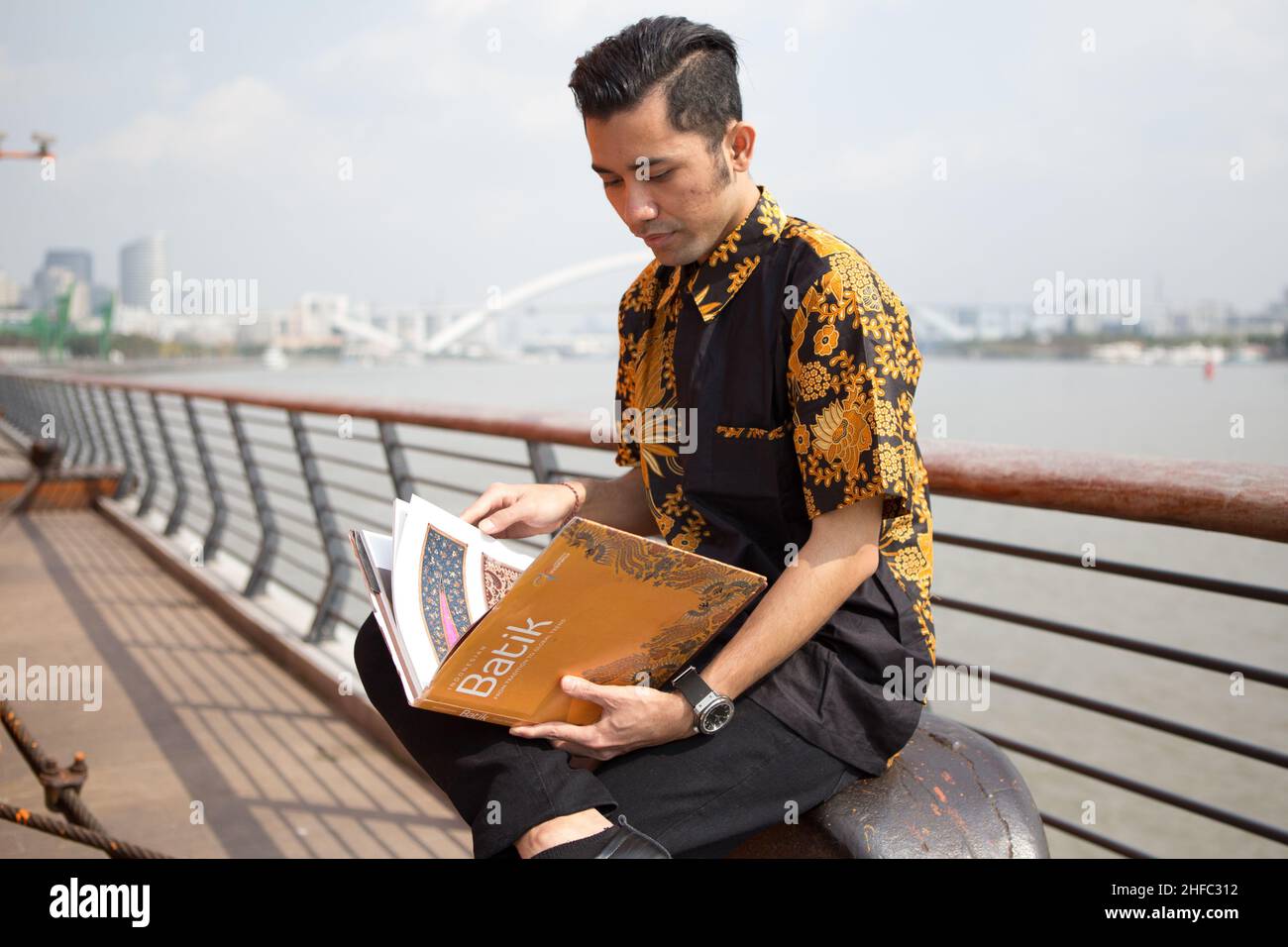 Un jeune modèle masculin habillé en indonésien Batik lit un livre intitulé Batik avec le fond de la rivière Huangpu et du pont de Lupu au long Museum Banque D'Images