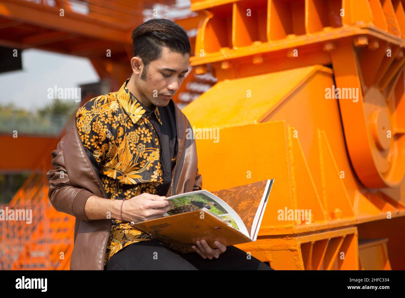 Un jeune modèle masculin vêtu de Batik indonésien lit un livre intitulé Batik et est assis sur une grande grue orange au long Museum, West Bund, Shanghai Banque D'Images