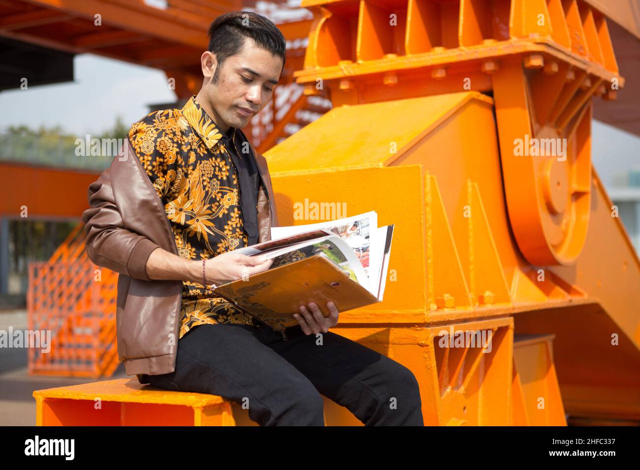 Un jeune modèle masculin vêtu de Batik indonésien lit un livre intitulé Batik et est assis sur une grande grue orange au long Museum, West Bund, Shanghai Banque D'Images