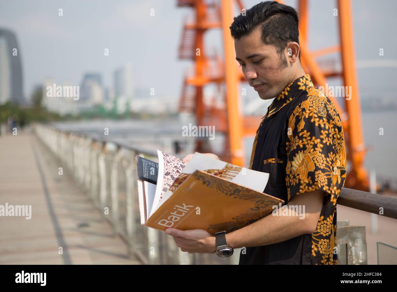 Un jeune modèle masculin habillé en indonésien Batik lit un livre sur Batik avec une grande grue orange en arrière-plan au long Museum, Shanghai Banque D'Images