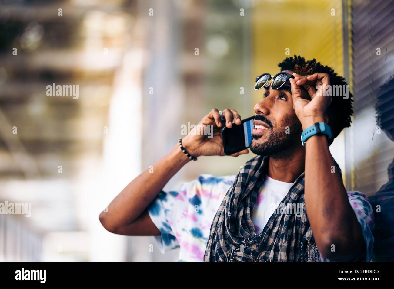 Gros plan d'un homme noir souriant appuyé contre un mur de verre avec son smartphone en profil. Retirer ses lunettes Banque D'Images