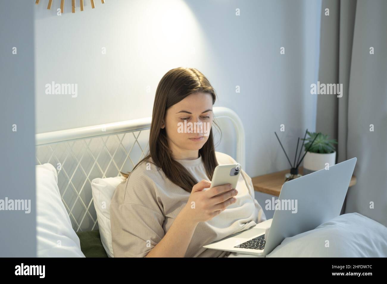Femme utilisant un ordinateur portable et surfant sur les réseaux sociaux sur un smartphone après avoir pris le lit Banque D'Images