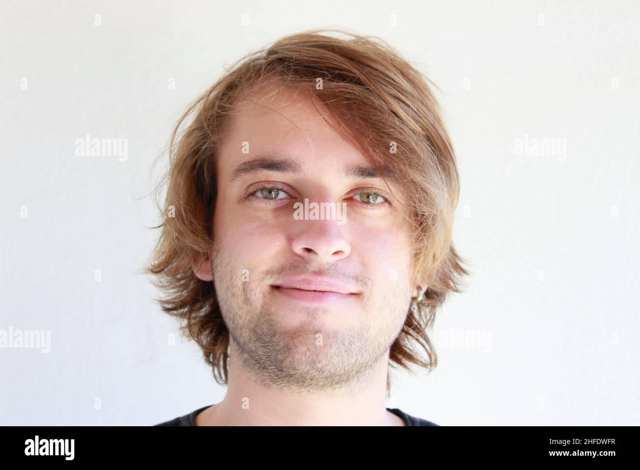 Jeune homme aux yeux verts souriant en franchise.Jeune homme aux longs cheveux blonds souriant isolé candidement sur fond blanc.Expressions du visage. Banque D'Images