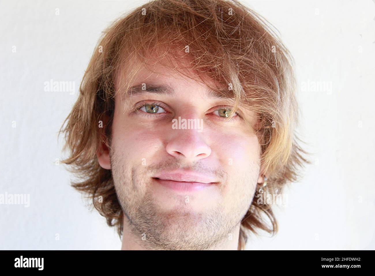 Jeune homme aux yeux verts souriant en franchise.Jeune homme aux longs cheveux blonds souriant isolé candidement sur fond blanc.Expressions du visage. Banque D'Images