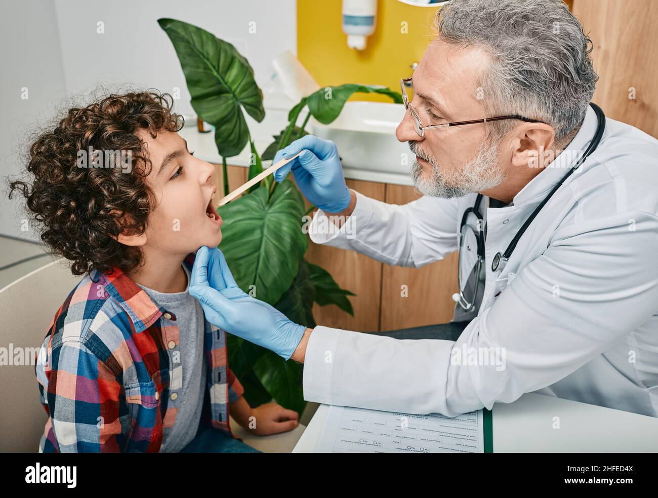 Médecin utilisant une spatule d'inspection pour examiner la gorge des garçons du moyen-Orient. Enfant mâle bouclé au rendez-vous du pédiatre Banque D'Images