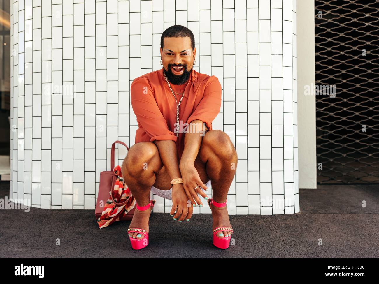 Faites glisser la reine en souriant à la caméra avec joie tout en vous squatant sur un fond blanc.Jeune homme non conforme portant du maquillage et des bijoux Banque D'Images