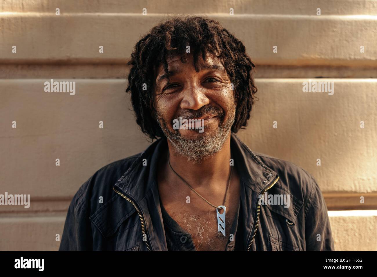 Homme discret souriant à la caméra en toute confiance tout en restant seul pendant la journée.Homme sans abri sûr de lui-même debout contre une brique wa Banque D'Images