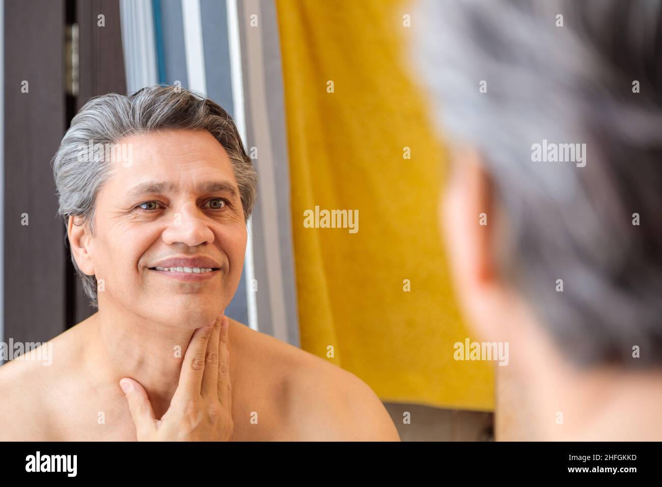 Un homme adulte aux cheveux gris sourit et regarde dans le miroir après le rasage. Banque D'Images