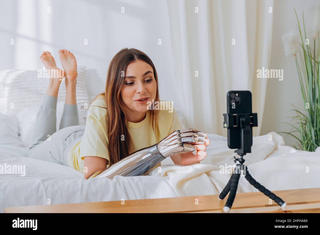 Une blogueuse bionique avec un bras prothétique dirige une émission en ligne, enregistre un problème ou communique avec des amis par appel vidéo tout en étant couché sur un lit dans des vêtements confortables de la maison par temps ensoleillé. Banque D'Images