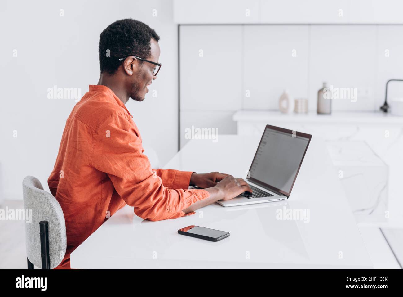 Un jeune homme afro-américain concentré en vêtements et lunettes orange travaille sur un ordinateur portable moderne dans un espace de travail confortable Banque D'Images