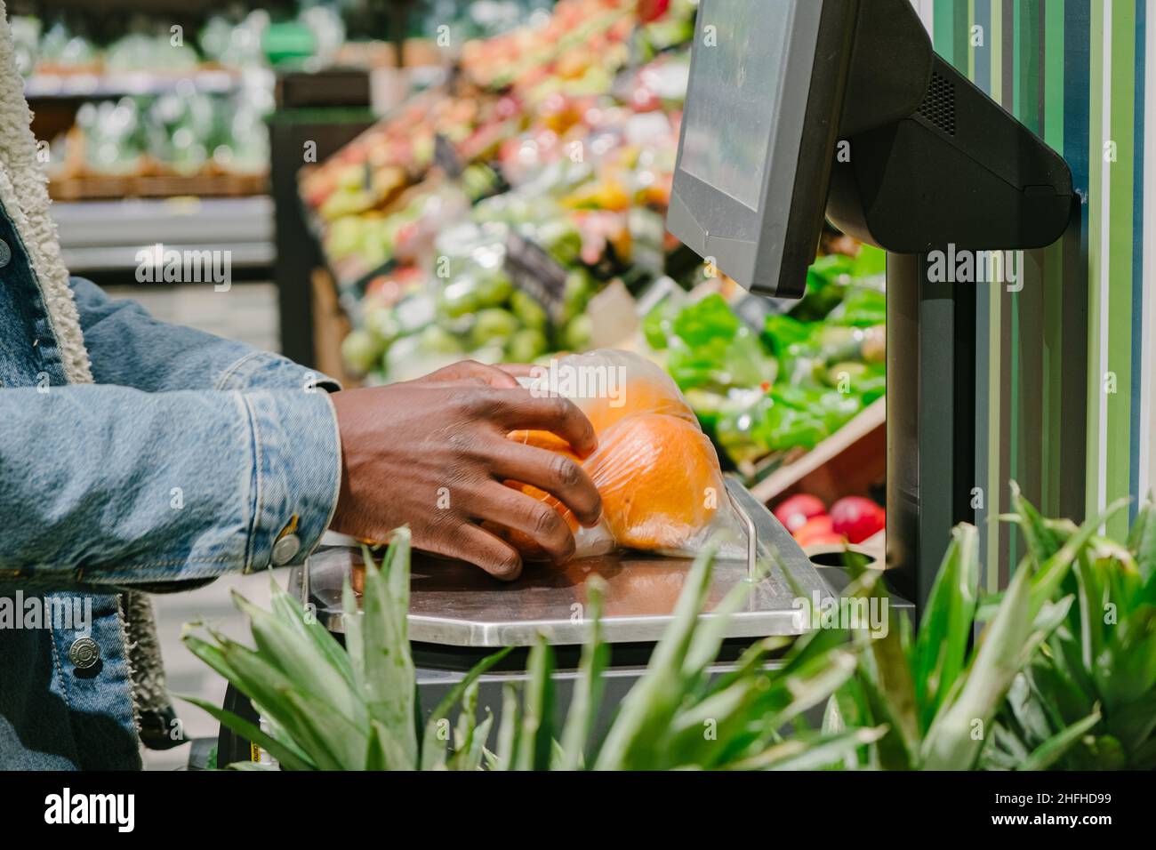 Un homme afro-américain élégant en veste chaude avec masque jetable pèse des oranges fraîches avec balance numérique dans un supermarché léger Banque D'Images