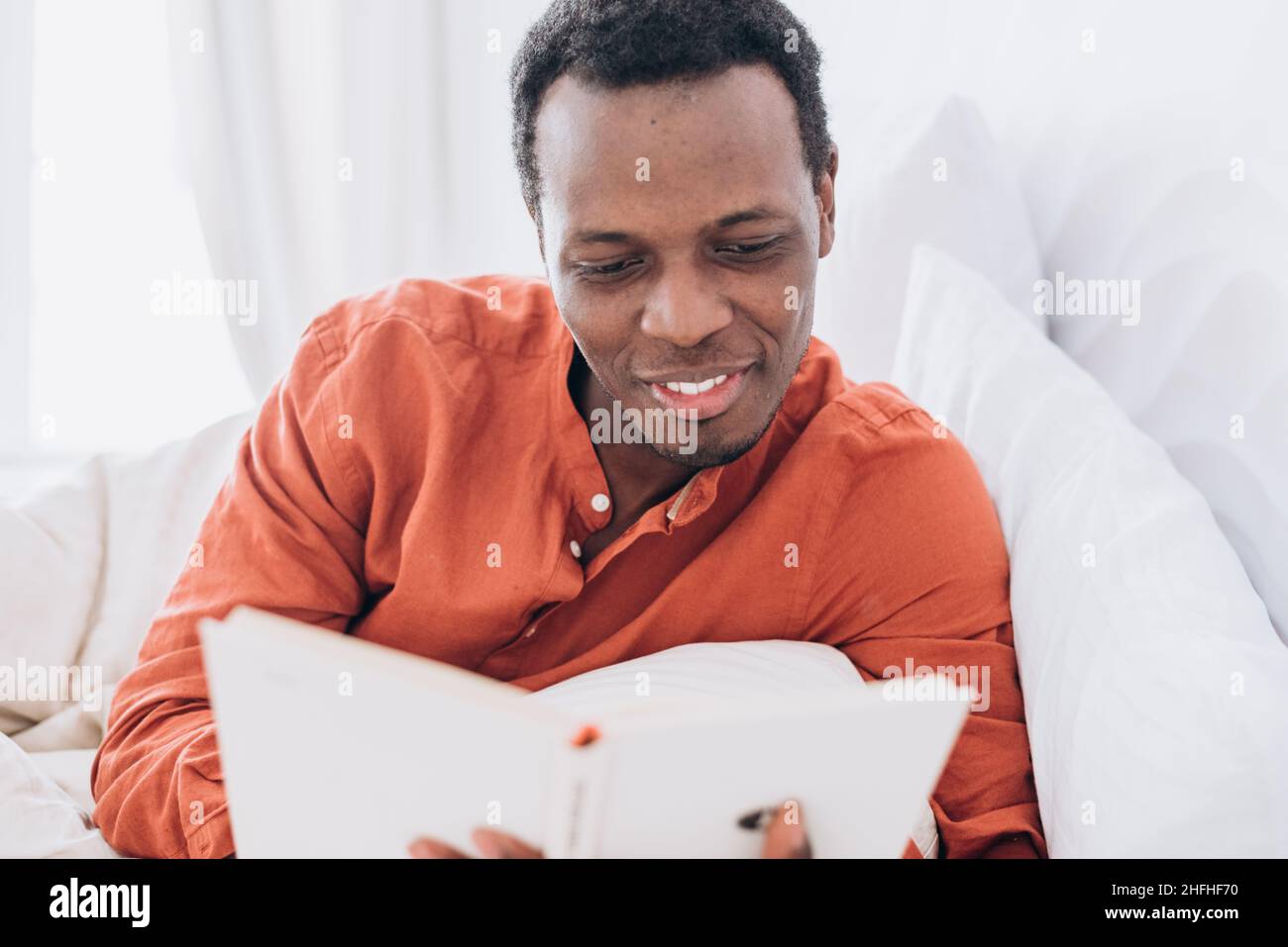 Beau jeune homme noir en pyjama lit un livre intéressant allongé dans un lit confortable avec oreillers doux et une couverture dans la chambre Banque D'Images