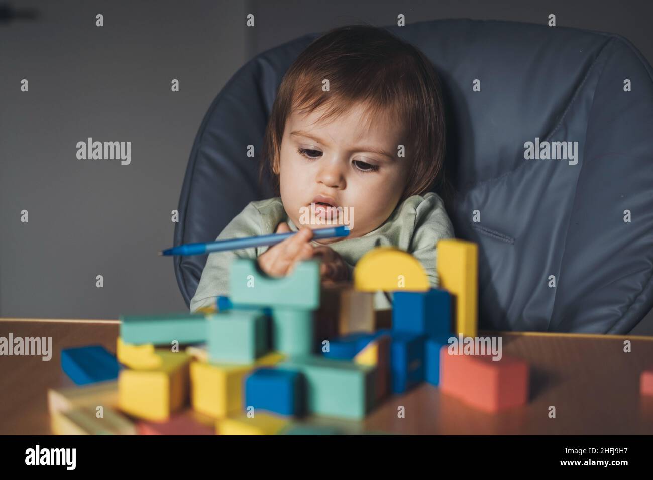 Bébé fille tenant un stylo dans ses mains tout en jouant avec des blocs de construction assis dans une chaise haute. Banque D'Images