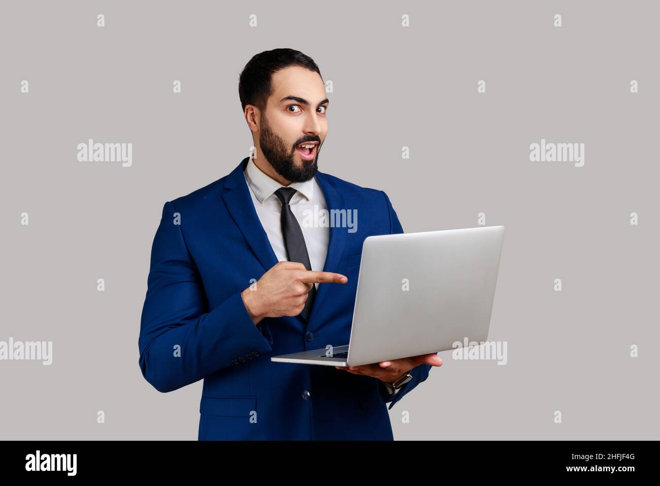 Homme barbu étonné pointant l'écran d'ordinateur portable et regardant la caméra avec l'expression choquée, la bouche ouverte, portant un costume de style officiel.Prise de vue en studio isolée sur fond gris. Banque D'Images