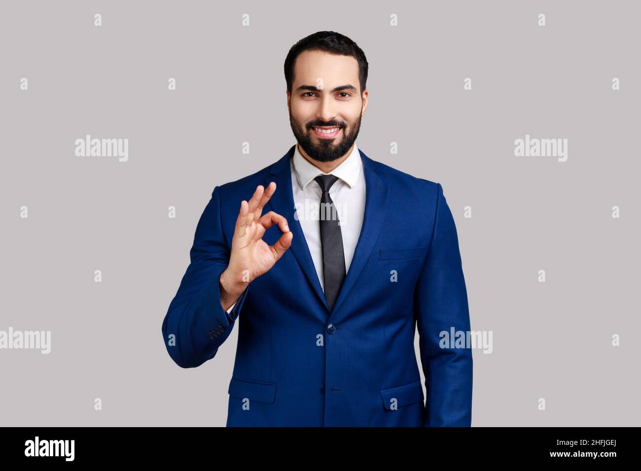 Portrait d'un homme barbu ravi debout, regardant l'appareil photo montrant le geste du signe OK, exprimant des émotions positives, portant un costume de style officiel.Prise de vue en studio isolée sur fond gris. Banque D'Images