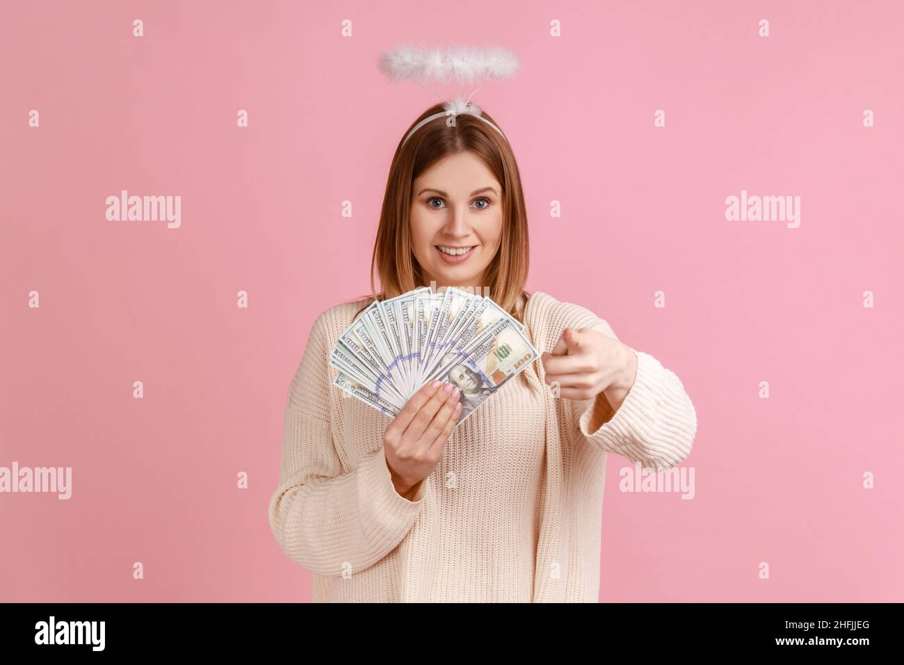 Portrait d'une femme gélique blonde satisfaite avec halo au-dessus de la tête pointant vers l'appareil photo et tenant un grand fan de billets de dollars, portant un chandail blanc.Studio d'intérieur isolé sur fond rose. Banque D'Images