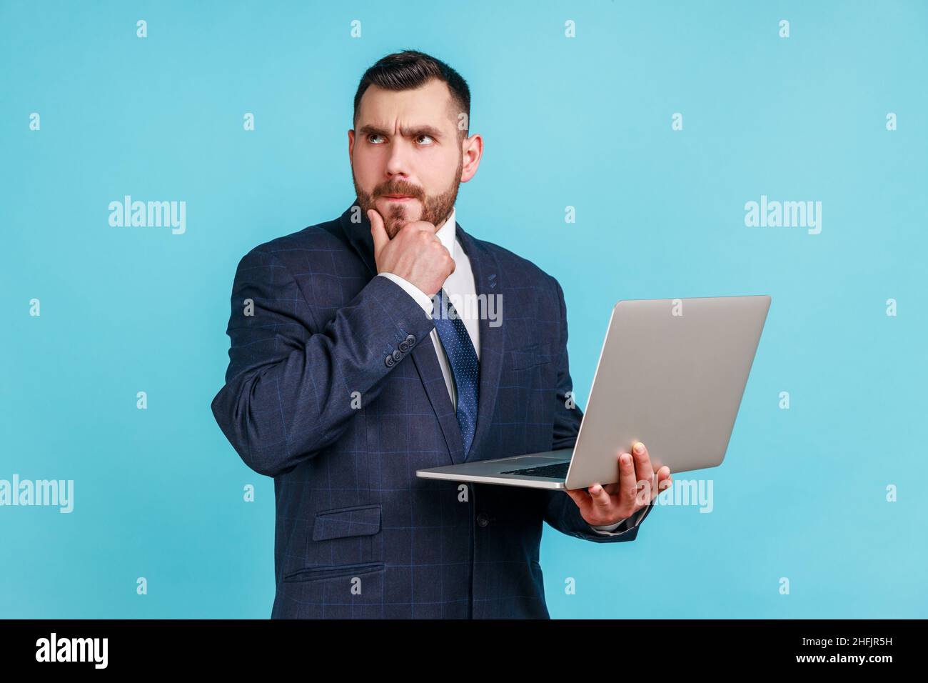 Portrait d'un homme d'affaires pensif élégant portant un costume sombre de style officiel tenant un ordinateur portable et pensant à un nouveau projet, tenant son menton.Studio d'intérieur isolé sur fond bleu. Banque D'Images