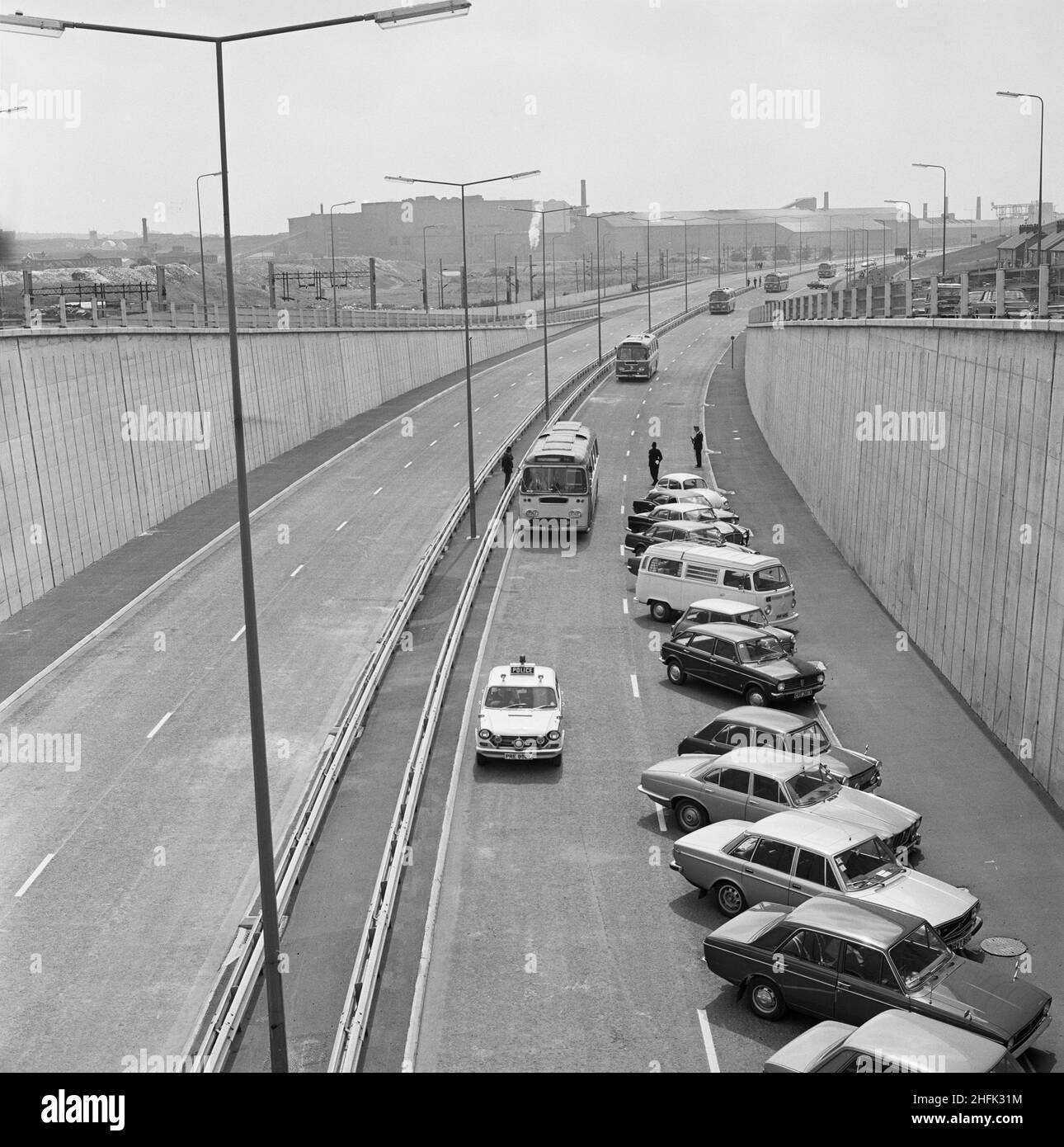 A500, Newcastle-Under-Lyme, Staffordshire, 27/06/1973.Une voiture de police conduisant une ligne d'autocars le long de la route Potteries D sur le A500 le jour d'ouverture officiel, passant devant une rangée de voitures garées sur la chaussée.Cette photographie fait partie d'un lot montrant la cérémonie d'ouverture d'un tronçon de 6km du chemin Potteries D ou du chemin Stoke D sur le A500, construit par la Direction des autoroutes de Laing. Banque D'Images