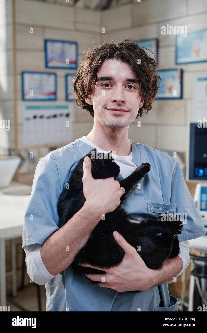 Portrait d'un jeune vétérinaire de sexe masculin en uniforme tenant un chat noir dans ses mains et regardant une caméra debout dans une clinique vétérinaire Banque D'Images