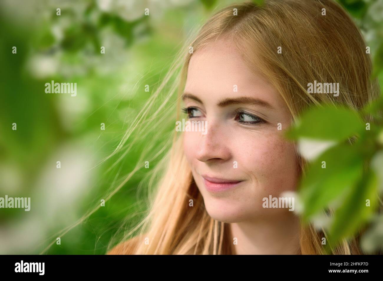 Portrait de belle femme souriante dans la nature encadrée par des branches floues avec des feuilles vertes fraîches et des fleurs blanches Banque D'Images