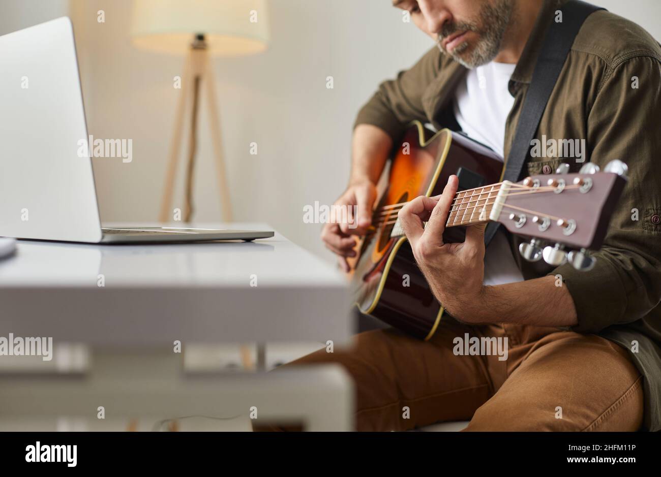 L'homme joue de la guitare acoustique à la maison devant le public en ligne ou donne des cours de musique à l'aide d'un ordinateur portable. Banque D'Images