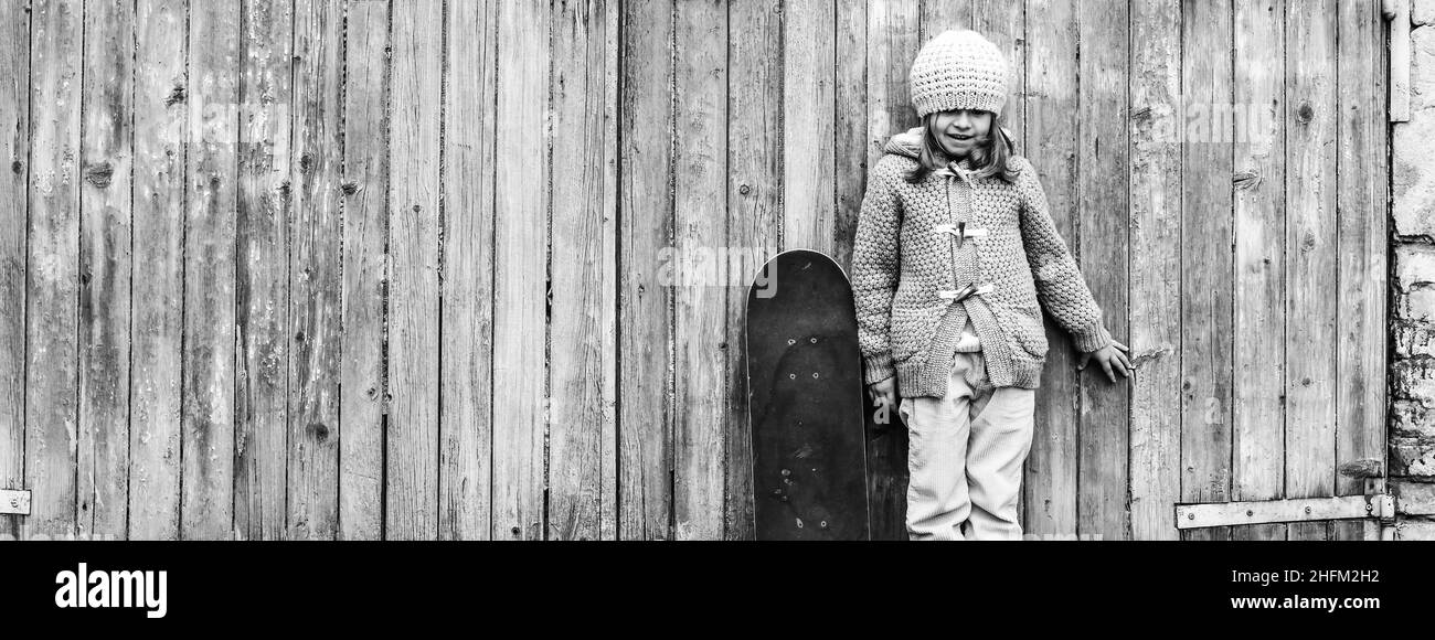 Bannière horizontale ou affiche supérieure avec adorable enfant fille en laine d'hiver vêtements avec planche à roulettes tout en se tenant contre fond en bois grunge - concept d'un Banque D'Images