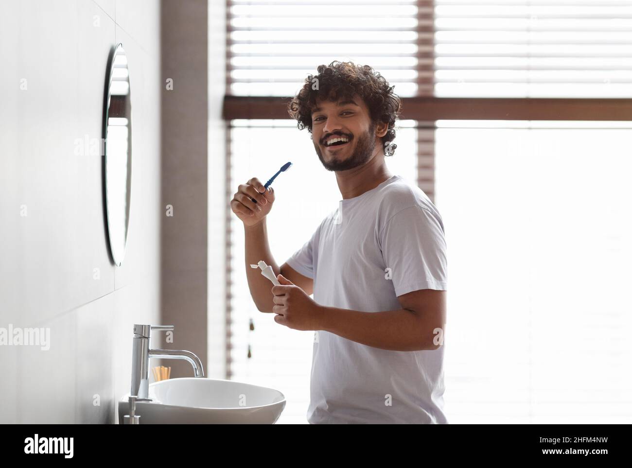 Concept de soins buccaux.Joyeux Indien barbu en t-shirt blanc se brossant les dents avec de la pâte, souriant à l'appareil photo dans la salle de bains Banque D'Images