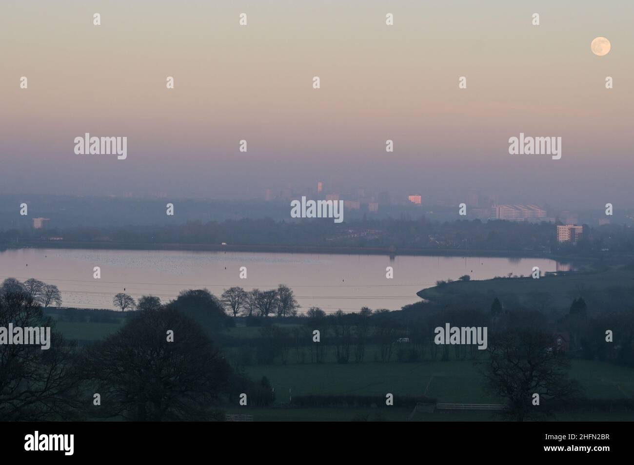 Frankley, Birmingham, Royaume-Uni. 17 janvier 2022. La première pleine lune de 2022, appelée Wolf Moon, s'élève au-dessus du réservoir Frankley au premier plan et de Birmingham qui est entouré d'une brume épaisse bloquant une grande partie de sa vue depuis les collines de Frankley, West Midlands. Le grand bâtiment blanc de l'hôpital Queen Elizabeth à Selly Oak est juste visible sous la lune. Photo par crédit : KStop Press Media/Alamy Live News Banque D'Images