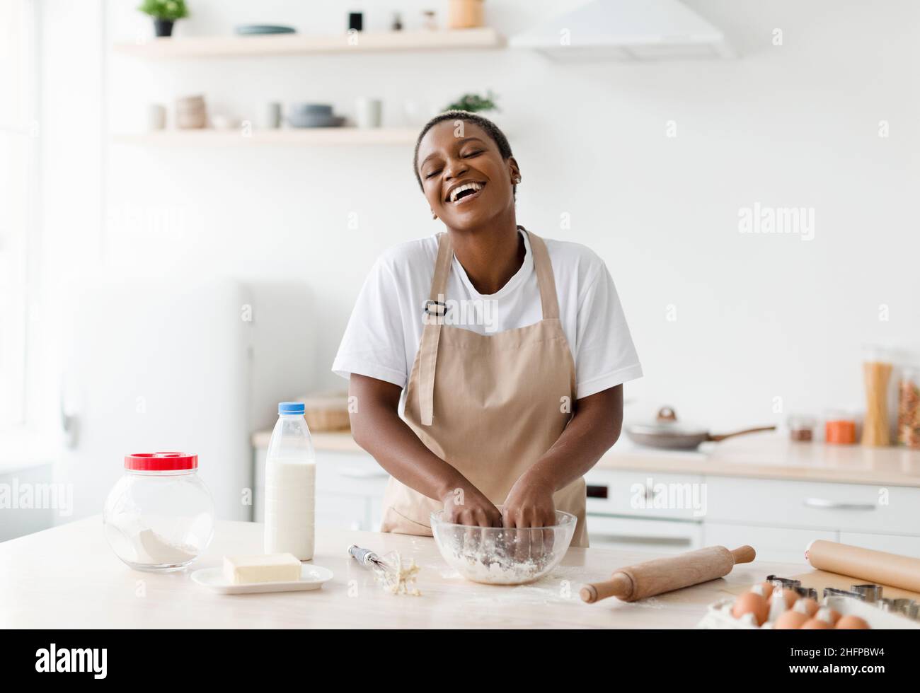 Heureux heureux rire jeune femme noire attrayante en tablier prépare la pâte pour la cuisson dans l'intérieur moderne de la cuisine Banque D'Images