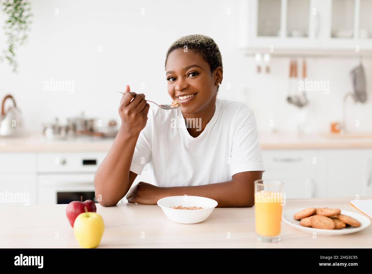 Souriant jeune femme noire attrayante mangeant porridge s'assoit à table avec des biscuits, jus et pommes Banque D'Images