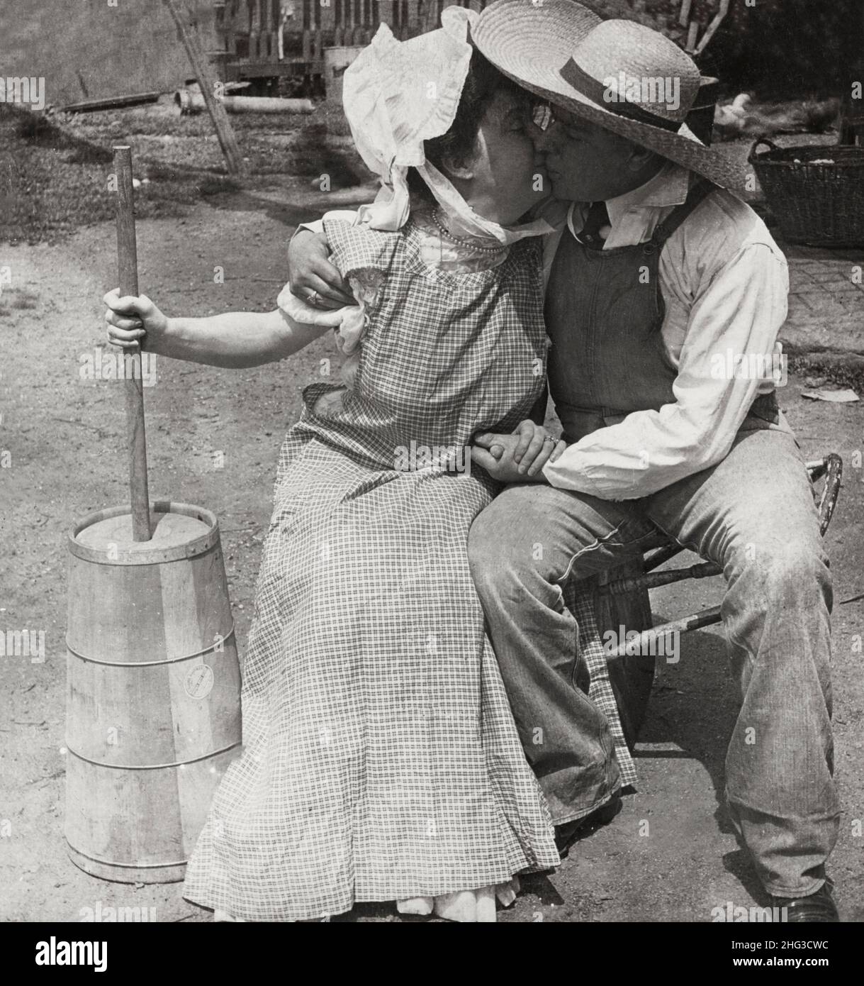 Photo d'amour vintage.Le binning, pas le muring.1906 le fermier et la femme de ménage embrassant; la femme avec le beurre churn. Banque D'Images
