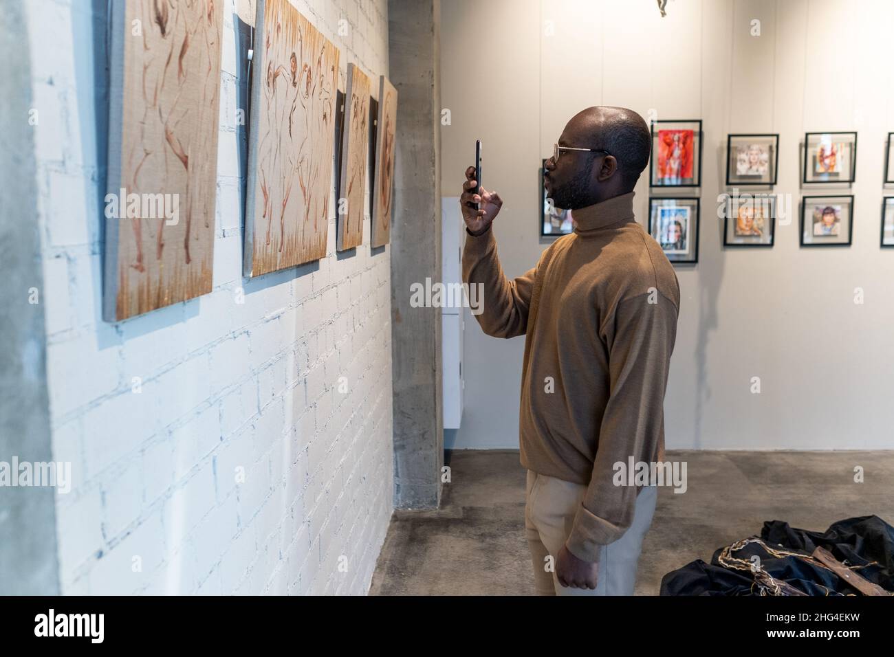 Jeune homme africain avec smartphone prenant des photos de peinture dans le musée tout en se tenant devant le mur avec la collection d'œuvres d'art Banque D'Images