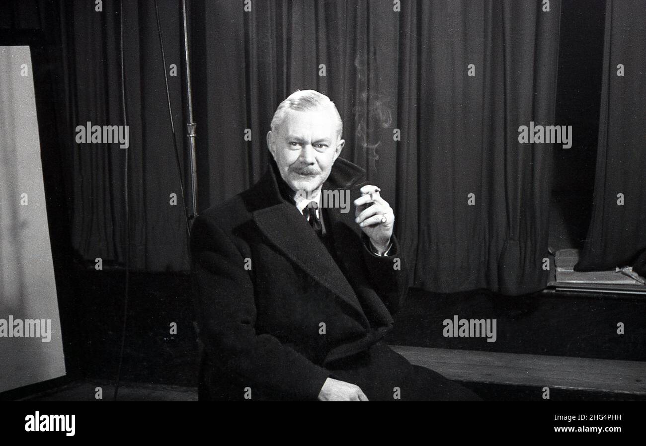 1950s, historique, un homme, peut-être un acteur dans les répétitions, assis dans son manteau par une scène et rideaux sur une pause, ayant une cigarette, Angleterre, Royaume-Uni. Banque D'Images