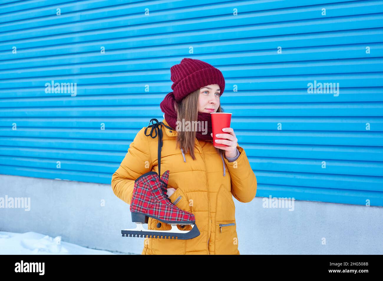 La femme skieuse, dans une veste jaune vif et chaude, boit du café à l'extérieur en hiver, près d'un fond bleu vif.Copier l'espace Banque D'Images