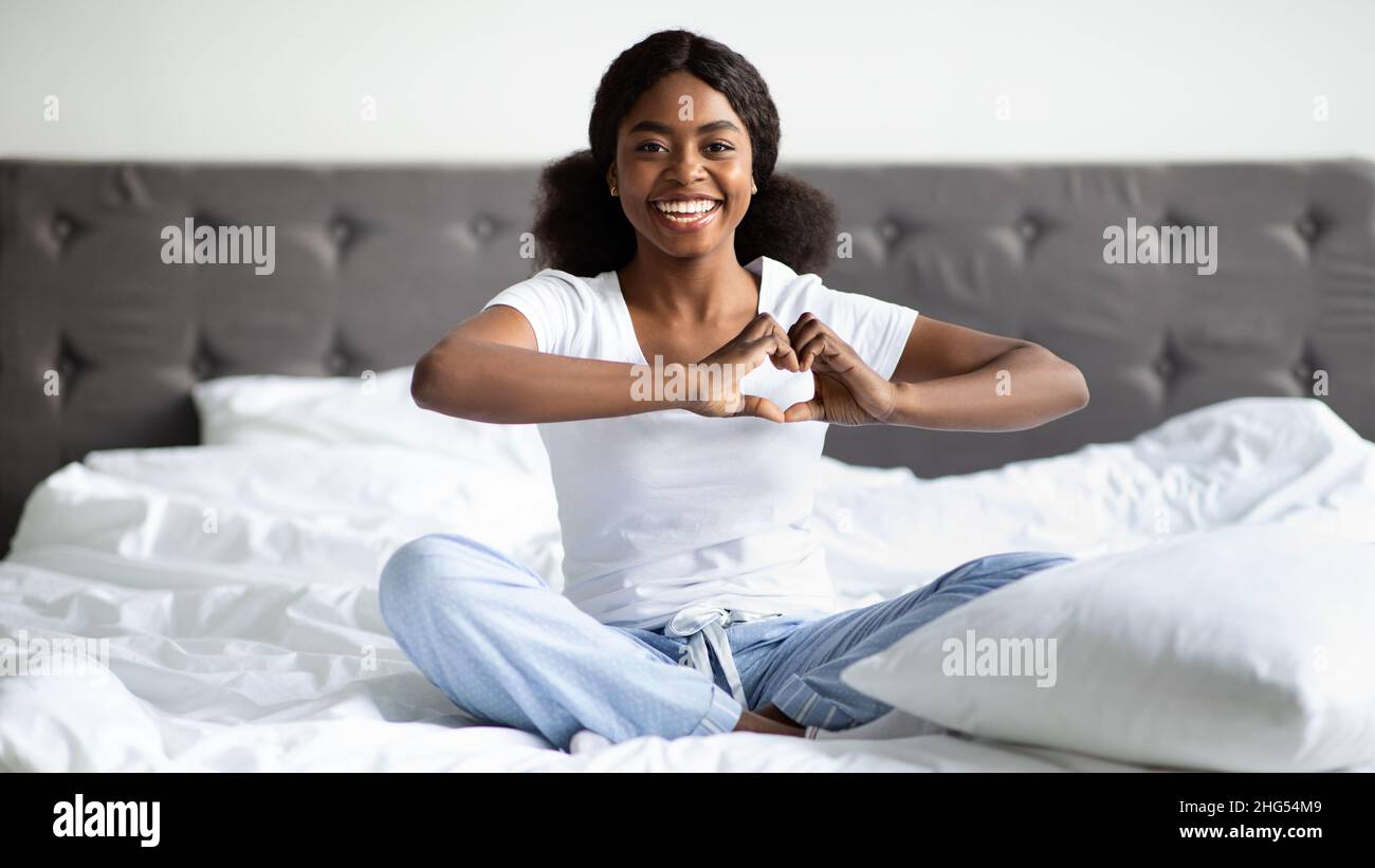 Femme noire positive en pyjama assise sur le lit, montrant le cœur Banque D'Images